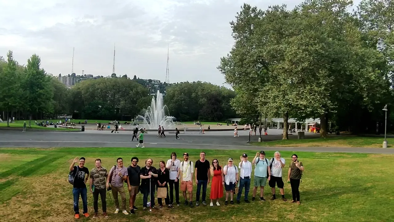 In the field in front of the International Fountain at the Seattle center. Picture taken from about 20 feet in the air from a drone - shows the Fair Worlds team lines up left to right waving at the camera and smiling.